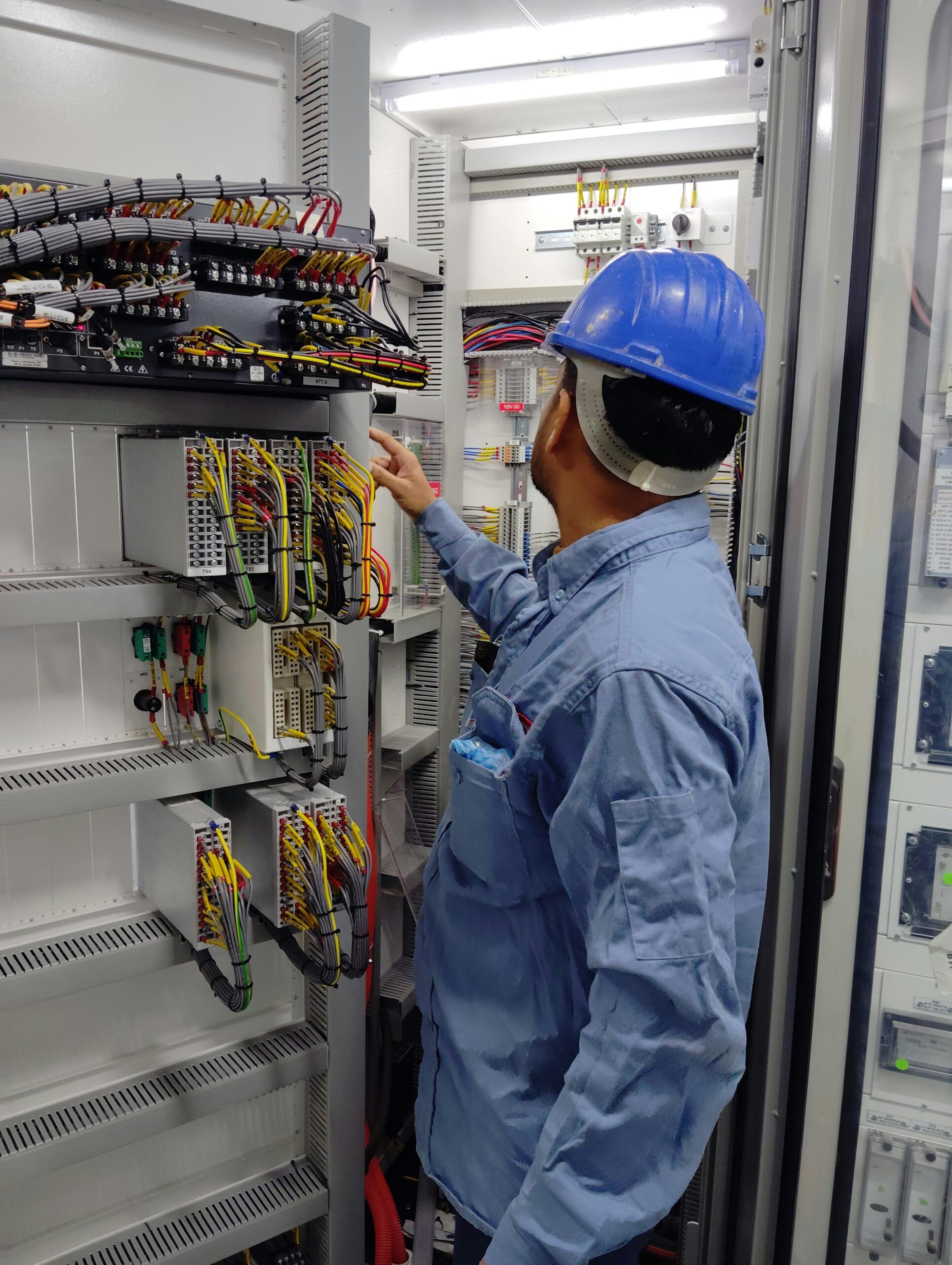 An electrician in a hard hat inspecting a control panel inside an industrial setting, ensuring safety and functionality.