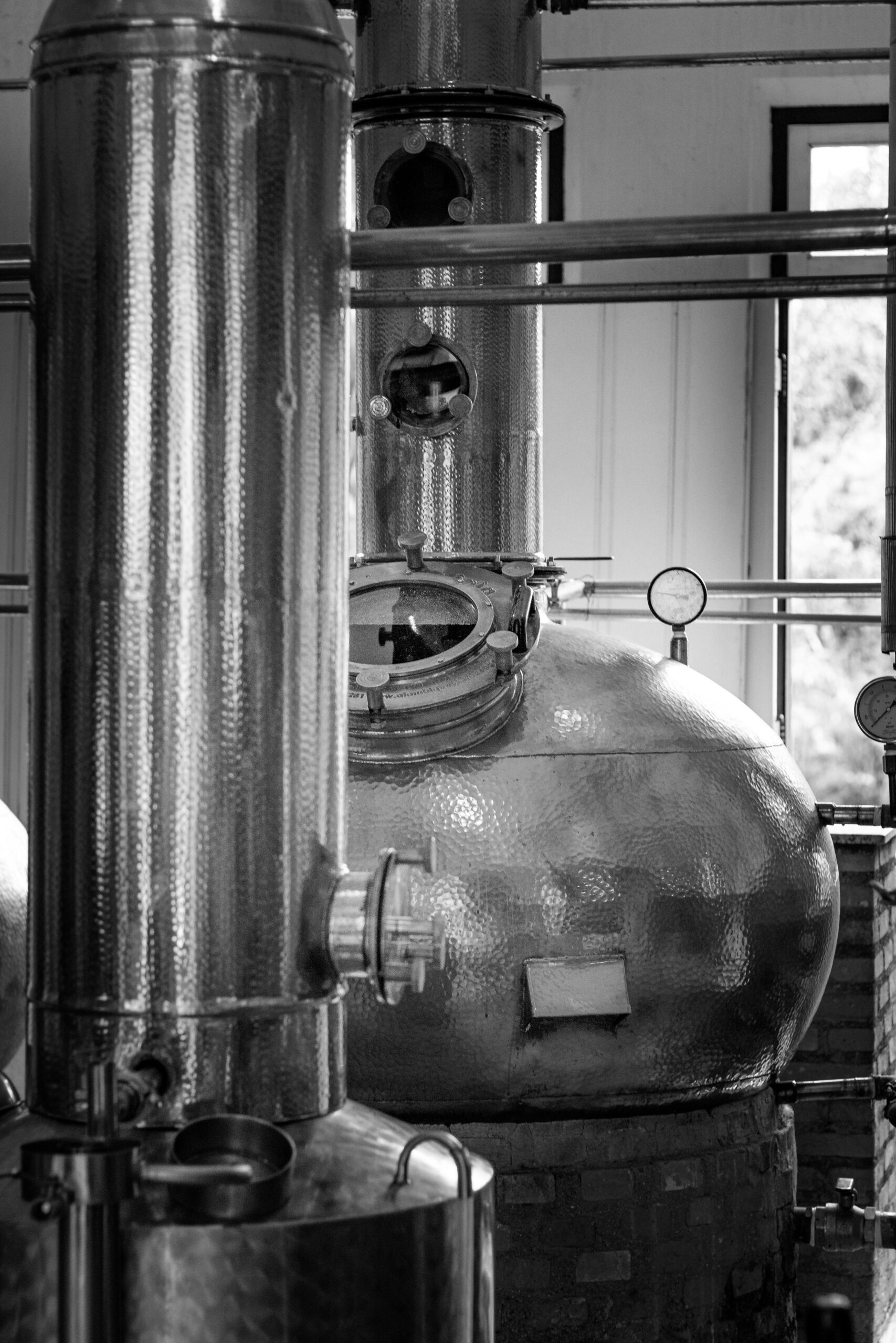 Black and white image of polished industrial distillery tanks and pipes indoors.